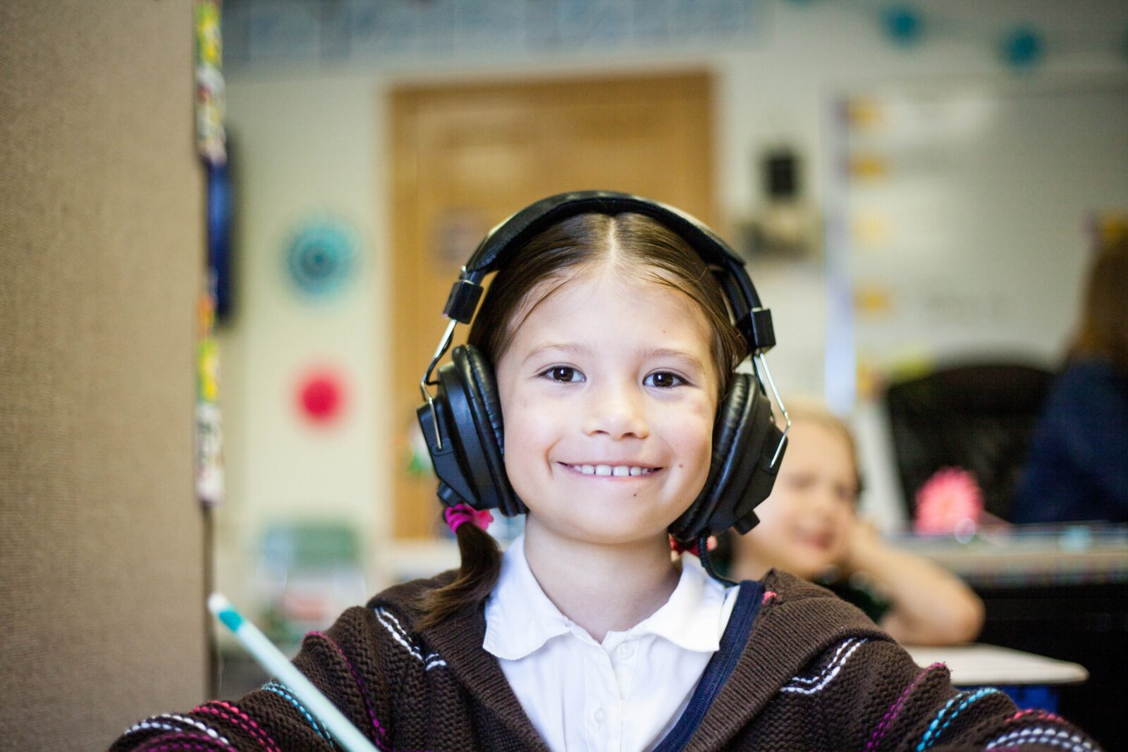 A young girl in a classroom wearing full-coverage headphones and smiling